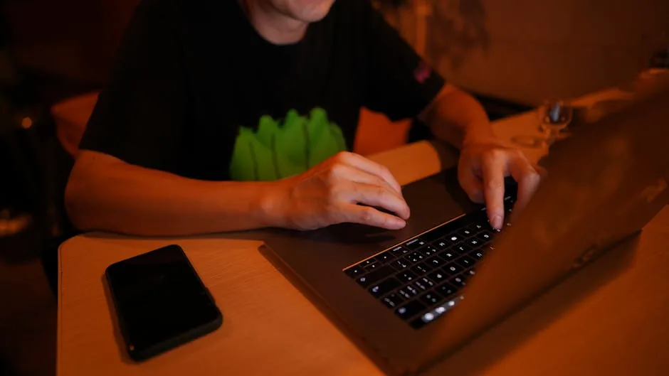 An adult man working at a desk on a laptop with a smartphone nearby, in a dimly lit home office.